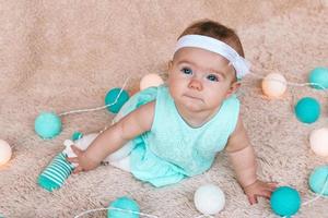 jolie petite fille en robe bleue est assise sur un plaid en peluche beige avec des lumières de noël avant le réveillon de noël. photo