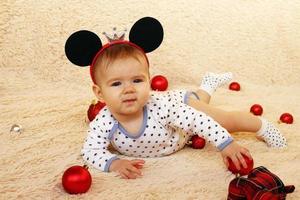 jolie petite fille aux oreilles de souris est allongée sur un plaid beige et joue avec des décorations de noël rouges. photo