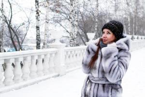 belle jeune femme aux cheveux rouges et aux yeux bleus en manteau de fausse fourrure grise avec capuche se promène dans le parc enneigé d'hiver. photo