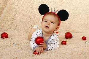 portrait d'une petite fille mignonne avec des oreilles de souris sur un plaid beige et des boules de noël brillantes rouges. photo