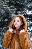 portrait d'une belle jeune femme aux cheveux roux et aux yeux bleus en manteau de fausse fourrure sur fond de parc enneigé d'hiver. photo