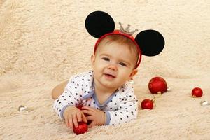 adorable petite fille souriante avec des oreilles de souris est allongée sur un plaid beige et joue avec des boules de noël rouges brillantes. photo