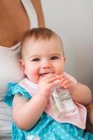 souriante petite fille caucasienne buvant de l'eau de la bouteille sur les genoux de la mère. l'enfant apprend à tenir une bouteille. photo