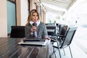 femme d'affaires boit du café dans un café avec un ordinateur portable sur la table regarde pensivement au loin photo