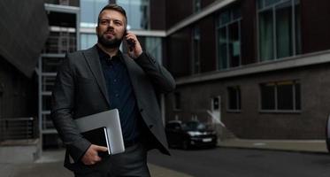 homme élégant adulte dans un costume d'affaires avec un ordinateur portable dans ses mains parle sur un téléphone mobile, concept d'une stratégie marketing réussie photo