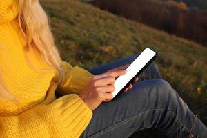 mains féminines avec téléphone, écran maquette blanc. femme blonde en pull jaune et jeans bleus à l'aide d'un smartphone, assise sur la colline à la lumière orange du coucher du soleil. image recadrée, sans visage, méconnaissable. photo