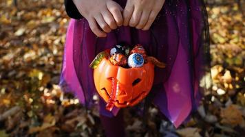 fille en robe violette de sorcière tient dans les mains un seau en plastique de citrouille orange avec des bonbons et des vers de gelée. forêt d'automne derrière. concept de truc ou de friandise d'halloween. pas de visage, méconnaissable, mise au point sélective. photo