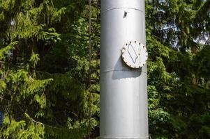 grande colonne de réservoir industriel en acier inoxydable brillant en fer avec une trappe ronde pliante, trou d'homme avec boulons, écrous et goujons sur fond d'arbres verts et de bois photo