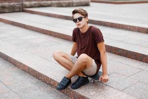 jeune homme en t-shirt avec des lunettes de soleil et un short assis sur une planche à roulettes. photo