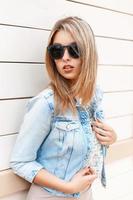 sexy belle fille élégante à lunettes de soleil et une veste en jean bleu debout près du mur en bois photo