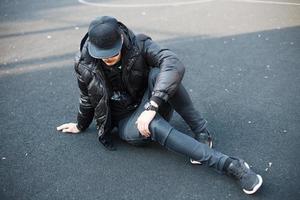 jeune homme élégant en vêtements noirs à la mode assis sur le trottoir photo