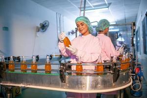 une travailleuse asiatique vérifie les bouteilles de jus de fruits sur la ligne de production de l'usine de boissons. le fabricant vérifie la qualité de l'industrie alimentaire. photo