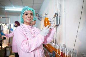 une travailleuse asiatique vérifie les bouteilles de jus de fruits sur la ligne de production de l'usine de boissons. le fabricant vérifie la qualité de l'industrie alimentaire. photo