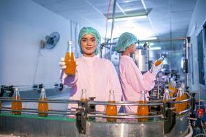 une travailleuse asiatique vérifie les bouteilles de jus de fruits sur la ligne de production de l'usine de boissons. le fabricant vérifie la qualité de l'industrie alimentaire. photo