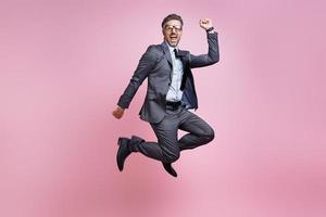 heureux homme mûr en costume complet sautant et gesticulant sur fond rose photo