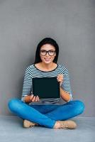 copier l'espace sur sa tablette. jolie jeune femme montrant sa tablette numérique et souriant assis sur le sol et sur fond gris photo