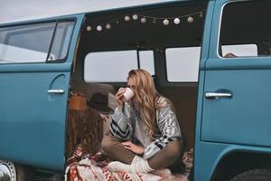 en dégustant une boisson chaude. jolie jeune femme buvant dans la tasse tout en étant assise à l'intérieur de la mini-fourgonnette bleue de style rétro photo