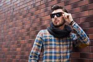 beau et avec style. beau jeune homme ajustant ses lunettes de soleil et regardant ailleurs tout en se tenant contre le mur de briques photo