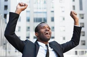 gagnant d'affaires. heureux jeune homme africain en tenue de soirée gardant les bras levés et exprimant sa positivité tout en se tenant à l'extérieur photo