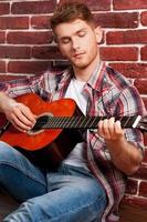 jeune et virtuose créatif. beau jeune homme jouant de la guitare acoustique tout en se penchant sur le mur de briques photo