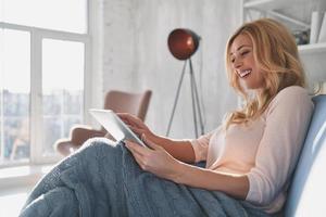 examinant sa nouvelle tablette. jolie jeune femme utilisant une tablette numérique et souriante assise sur le canapé à la maison photo
