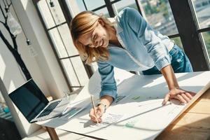 jolie jeune femme écrivant quelque chose sur le plan et souriant tout en travaillant au bureau photo