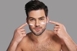 prendre soin de sa peau. beau jeune homme souriant appliquant une crème hydratante et regardant la caméra en se tenant debout sur fond gris photo