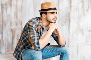 attentionné et romantique. beau jeune homme portant un chapeau et regardant ailleurs assis contre le mur en bois photo