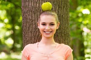 en harmonie avec la nature. belle jeune femme portant une pomme sur la tête et regardant la caméra avec le sourire tout en se penchant sur l'arbre dans un parc photo