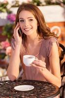 parler avec un ami. jolie jeune femme parlant au téléphone mobile et souriant tout en étant assis au café en plein air photo