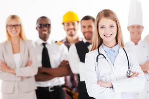 Quand je serai grand, je serai médecin. belle jeune femme médecin gardant les bras croisés et souriant tout en groupe de personnes dans différentes professions debout en arrière-plan photo