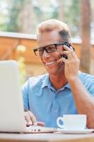 appréciant son travail à l'extérieur. heureux homme mûr travaillant sur un ordinateur portable et parlant au téléphone portable tout en étant assis à la table à l'extérieur avec la maison en arrière-plan photo