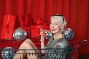 Senior élégante femme aux cheveux gris en lunettes de soleil et robe argentée assise dans le chariot de supermarché à la fête, buvant du vin sur fond de rideaux rouges. fête, disco, célébration, concept d'âge senior photo