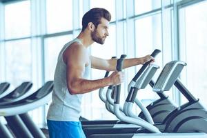 travailler pour le meilleur corps. vue latérale d'un jeune bel homme en tenue de sport regardant loin tout en travaillant sur un stepper à la salle de sport photo