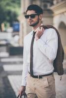 prêt pour un rendez-vous. beau jeune homme à lunettes de soleil tenant une veste et regardant loin tout en marchant à l'extérieur photo