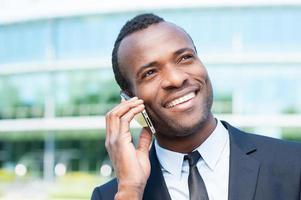 parler d'affaires. confiant jeune homme africain en tenues de soirée parlant au téléphone mobile et souriant tout en se tenant à l'extérieur photo