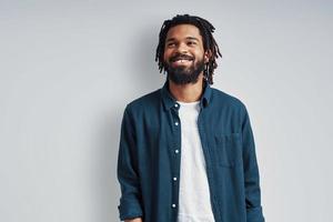 charmant jeune homme africain en tenue décontractée souriant et regardant ailleurs en se tenant debout sur fond gris photo