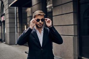 son style est parfait. beau jeune homme en tenue décontractée intelligente ajustant ses lunettes tout en marchant dans la rue de la ville photo