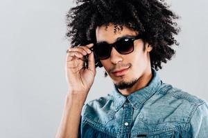 regarde-moi de plus près. portrait d'un beau jeune homme africain regardant la caméra et ajustant ses lunettes de soleil en se tenant debout sur fond gris photo