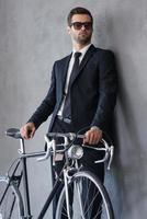 parfait avec son nouveau vélo. confiant jeune homme d'affaires regardant loin tout en se tenant debout avec son vélo sur fond gris photo
