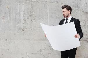 plan pour un nouveau projet. beau jeune homme examinant le plan en se tenant debout à l'extérieur et contre le mur de béton photo