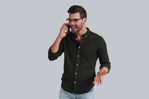 en contact avec les clients. beau jeune homme à lunettes parlant au téléphone intelligent et souriant en se tenant debout sur fond gris photo