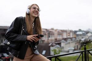 une jeune étudiante écoute de la musique agréable sur fond de paysage urbain dans des écouteurs avec un téléphone dans les mains photo