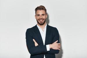 si beau. beau jeune homme en costume complet gardant les bras croisés et regardant la caméra avec le sourire en se tenant debout sur fond gris photo