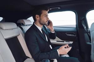 toujours disponible. beau jeune homme en costume complet parlant au téléphone intelligent alors qu'il était assis dans la voiture photo