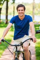 profitant de sa promenade dans le parc. beau jeune homme à vélo dans le parc et souriant à la caméra photo
