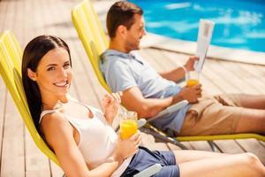 profiter de l'orange fraîche au bord de la piscine. vue latérale d'un beau jeune couple assis sur les chaises longues au bord de la piscine pendant qu'une femme regarde la caméra et sourit photo