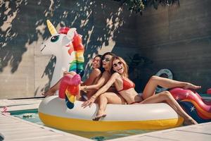 jolies jeunes femmes en maillot de bain souriant tout en flottant sur une licorne gonflable dans la piscine en plein air photo