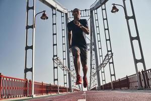 en plein forme. toute la longueur du jeune homme africain en vêtements de sport faisant de l'exercice en faisant du jogging sur le pont à l'extérieur photo