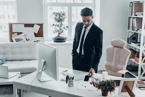 plus d'appels téléphoniques pour aujourd'hui. beau jeune homme en costume complet posant son téléphone intelligent sur le bureau tout en se tenant au bureau photo